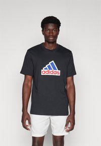 Černé bavlněné tričko s velkým červeným a modrým logem Adidas na hrudi. Krátké rukávy, kulatý výstřih, standardní střih.