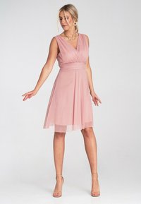 Rosa ärmelloses Kleid mit V-Ausschnitt, geraffter Taille und fließendem Rock. Weicher Stoff mit transparentem Überzug, ergänzt durch schlichten Schmuck.
