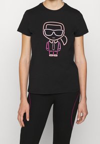 Camiseta negra con una figura de dibujos animados con gafas y una coleta, contornada en rosa y púrpura. Tela suave, mangas cortas, diseño ajustado.