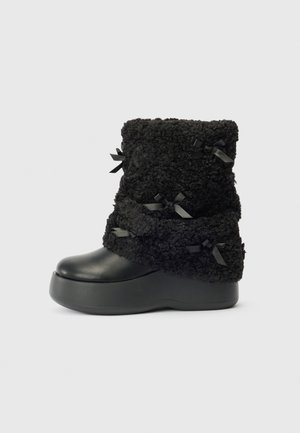 Botte chelsea noire à talon plat avec une tige en fausse fourrure texturée et de petits nœuds en satin décoratifs, vue de côté sur fond uni.