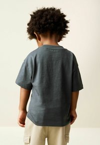 Sötétszürke rövid ujjú póló bordázott nyakkal, pamut anyagból, kényelmes szabással, világos bézs nadrággal párosítva.