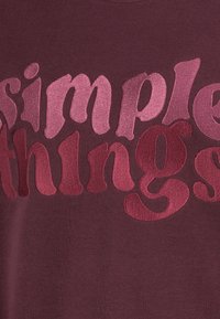 Burgundy sweatshirt featuring bold, textured lettering in varying shades of pink spelling "simple things." Soft fabric, relaxed fit.
