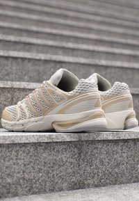 Paar beige und weiße Mesh-Sportschuhe auf grauen Betontreppen mit strukturierten Details an Sohlen und Seiten.
