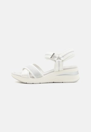 White sandals with fabric straps, an adjustable ankle strap, and a cushioned insole. The sole has a slight wedge and reflective accent.
