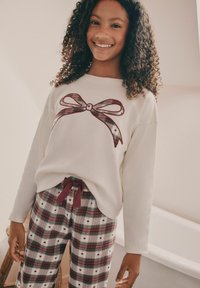 Vitt långärmat topp med en grafisk rutig rosett, kombinerat med rutig pyjamasbyxa med hjärtan i rött och grönt, fästa med ett vinrött snöre.