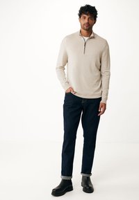 Maglione beige chiaro a zip metà con una texture morbida, abbinato a jeans blu scuro arrotolati e stivaletti neri. Design semplice e vestibilità rilassata.