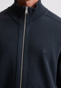 Marineblauer Strickjacke mit hohem Kragen, strukturiertem Stoff und silbernem Reißverschluss. Mit dezentem Branding auf der Brust.