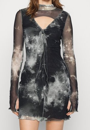 La femme porte une robe noire cintrée avec de longues manches transparentes et un imprimé galaxie, avec un nœud à l'avant et une petite découpe au niveau de la poitrine.