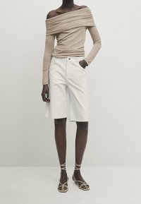 Beige schulterfreies Langarmoberteil kombiniert mit weißen knielangen Shorts und braunen Riemchensandalen. Glatter Stoff mit einer figurbetonten Silhouette.