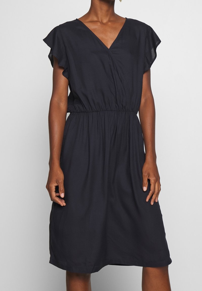 Femme portant une robe noire longueur genou avec des manches volantes et une taille froncée, debout devant un fond clair uni.