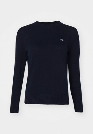 Mørkeblå strikkegenser med rund hals, lange ermer og ribbestrikket kant. Har en liten brodert logo på brystet.