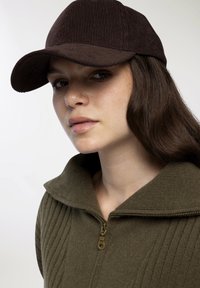 Brązowy sztruksowy czapka baseballowa w połączeniu z zielonym swetrem zapinanym na zamek z szerokim kołnierzem i mosiężnym detalem zamka.