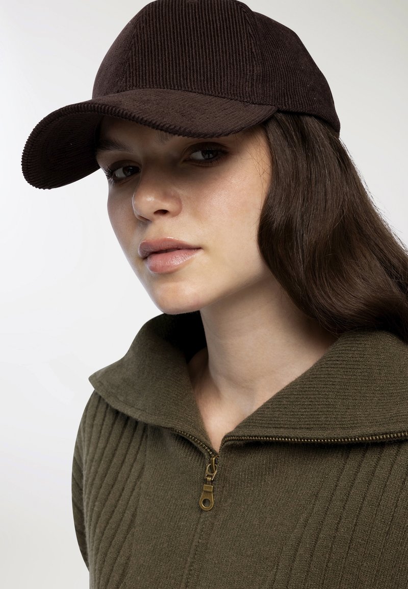 Brązowy sztruksowy czapka baseballowa w połączeniu z zielonym swetrem zapinanym na zamek z szerokim kołnierzem i mosiężnym detalem zamka.