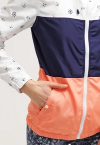 Farverig jakke med en hvid øverste del med anker mønstre, en navy midterdel og en koral nederste del. Inkluderer en front lynlås og lommer.