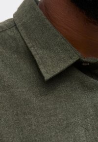 Gros plan sur un col de chemise en tissu vert foncé porté par une personne à la peau foncée et aux courts poils faciaux.