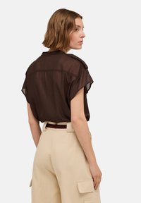 Braune, durchsichtige Bluse mit kurzen Ärmeln und Knopfdetails an der Schulter, kombiniert mit hochgeschnittenen, beigen Cargo-Hosen mit seitlichen Taschen.