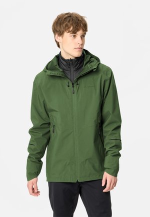 Jeune homme portant une veste de pluie à capuche verte avec une doublure noire, debout devant un fond clair uni, regardant vers la droite.