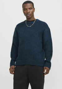 Navyblaue gestrickte Pullover mit strukturierter Oberfläche, Rundhalskragen und gerippten Bündchen, kombiniert mit schwarzen Hosen. Schlichter silberner Kettenakzent.