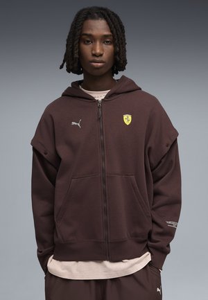 Ung person med korte dreadlocks iført en brun Puma-lynlås-hættetrøje med et gult Ferrari-logo, med hænderne i lommerne.