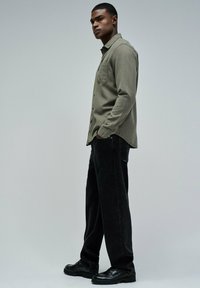 Joven de pie de lado con una camisa de botones verde oliva, pantalones negros y zapatos de cuero negros frente a un fondo gris liso.