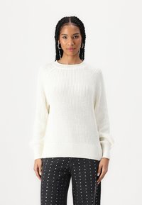 Gemütlicher weißer Strickpullover mit gerippten Bündchen, Rundhalsausschnitt und strukturiertem Muster; kombiniert mit schwarzen Jeans, die mit silbernen Nieten verziert sind.