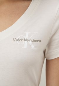 Bežinė V formos marškinėlių apykaklė, pagaminta iš minkštos medvilnės. Savo dizainu turi baltą "CK" logotipą ir žodžius "Calvin Klein Jeans" pilka spalva. Trumpomis rankovėmis, sklandžios tekstūros.