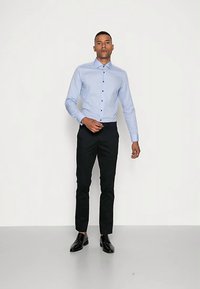 Lichtblauw overhemd met lange mouwen en donkere knopen, gecombineerd met strakke zwarte broek en zwarte schoenen, tegen een neutrale achtergrond.