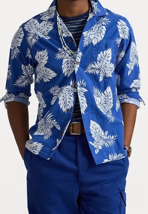 Man met een blauw overhemd met knopen en een wit tropisch bladmotief, een marineblauw gestreept ondershirt, blauwe broek, riem, kralenhalsketting en polshorloge, handen in de zakken.