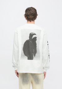 Hvid langærmet t-shirt med en stor sort-hvid grafik af en figur med hætte på bagsiden. Blødt bomuldsmateriale.