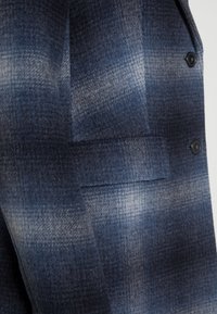 Blauw en grijs geruite wol stof van een jas, met een voorzak en twee zwarte knopen op een lichte achtergrond.