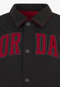Svart jakke med rød og grå brodert "JORDAN"-tekst, svarte knapper og rød mesh-fôr. Har en klassisk krage-design og sportslige detaljer.