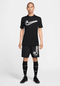 Černé sportovní oblečení zahrnující tričko s bílým logem Nike, odpovídající šortky, ponožky po kolena a černé sportovní boty.