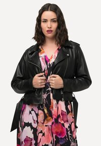 Giacca da motociclista in pelle nera con zip e dettagli in bottone, indossata sopra un vestito floreale colorato con tonalità di rosa e viola.