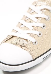 Zapatillas doradas con un acabado metálico texturizado, cordones blancos y una punta de goma blanca. Incluye ojales plateados y una suela negra.