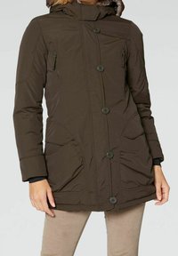 Brun vinterjacka med knäppning, dragkedjefickor på bröstet, stora fickor med klaff fram och en huva med pälsinslag, bärs över beige byxor.