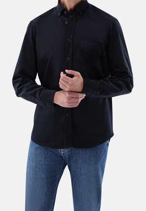 Homme portant une chemise noire à manches longues boutonnée et un jean bleu, ajustant une manchette avec la main opposée.