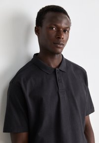 Cămașă polo din bumbac negru, cu guler, placket cu două nasturi și detaliu subtil al logo-ului. Textură netedă și mâneci scurte.