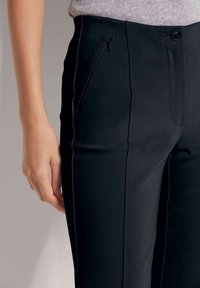 Zwarte broek van een gladde stof, met een rechte pasvorm, voorzakken met ritsen en een dubbele sluiting met een knop op de taille.