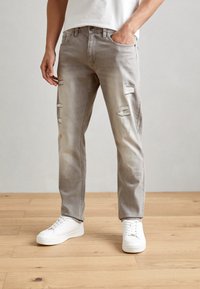 Gray distressed jeans with a faded finish, featuring multiple rips. Worn with white sneakers on a wooden floor, minimal background.
