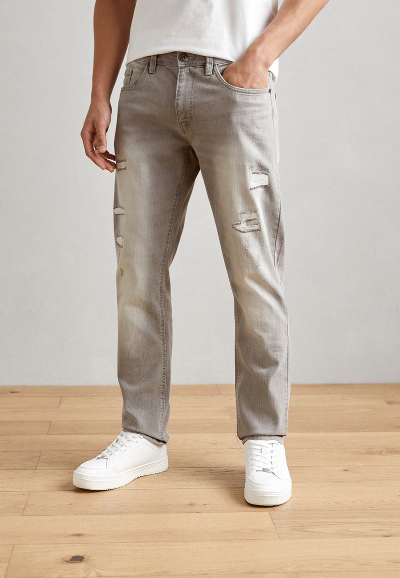 Gray distressed jeans with a faded finish, featuring multiple rips. Worn with white sneakers on a wooden floor, minimal background.