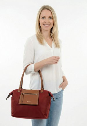 Blondide juustega naine, kes kannab valget pluusi ja teksapükse, hoiab suurt tumegranaatpunast käekotti pruunide nahast käepidemete ja esitaskuga.