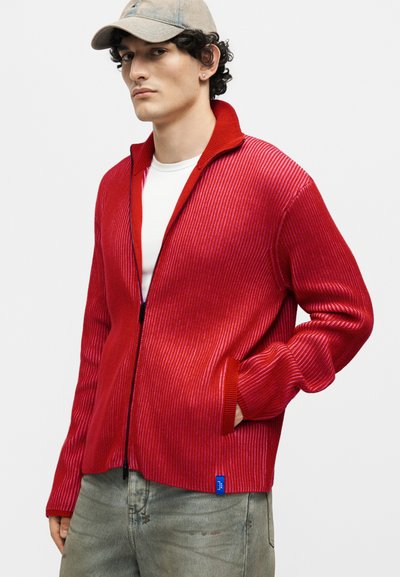 Νέος άνδρας που φοράει κόκκινο ριμπ πλεκτό πουλόβερ με φερμουάρ πάνω από λευκό πουκάμισο, γκρι τζιν και μπεζ καπέλο μπέιζμπολ, με το ένα χέρι στην τσέπη.