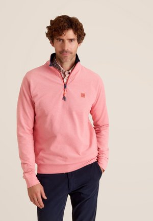 Pull-over rose clair avec un zip quart, col bleu marine et un motif texturé subtil. Présente un petit logo sur la poitrine. Associé à un pantalon bleu marine.