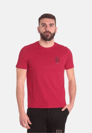 Uomo con barba che indossa una maglietta rossa a maniche corte con piccolo testo nero sul petto e pantaloni neri, in piedi davanti a uno sfondo chiaro e uniforme.