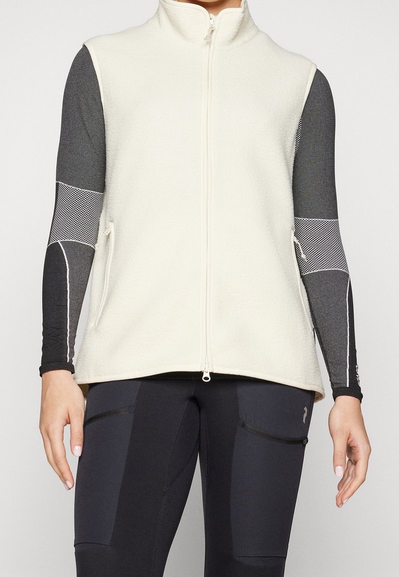 Personne portant une veste polaire blanche sans manches zippée sur un haut de sport noir et gris à manches longues à motifs et des leggings noirs.