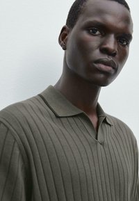 Olive green, short-sleeved polo shirt with a ribbed texture and a slight v-neck, featuring a dark collar and subtle vertical stripes.