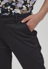 Svarta skräddarsydda byxor med en mjuk textur, utrustade med sidofickor och en åtsittande design, kombinerade med en blommig blus i lila och beige nyanser.