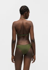 Mujer con el cabello trenzado, vistiendo un bikini verde oliva, de pie de espaldas contra un fondo gris claro y liso.