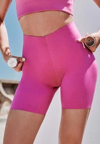 Femme en tenue de sport rose tenant des haltères de 1 kg, se concentrant sur la zone abdominale et le haut des jambes en plein air.