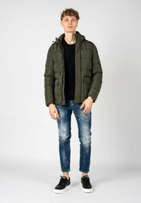 Olijfgroene pufferjack met capuchon, voorzien van gestikte delen, gecombineerd met versleten blauwe spijkerbroeken en zwarte sneakers.